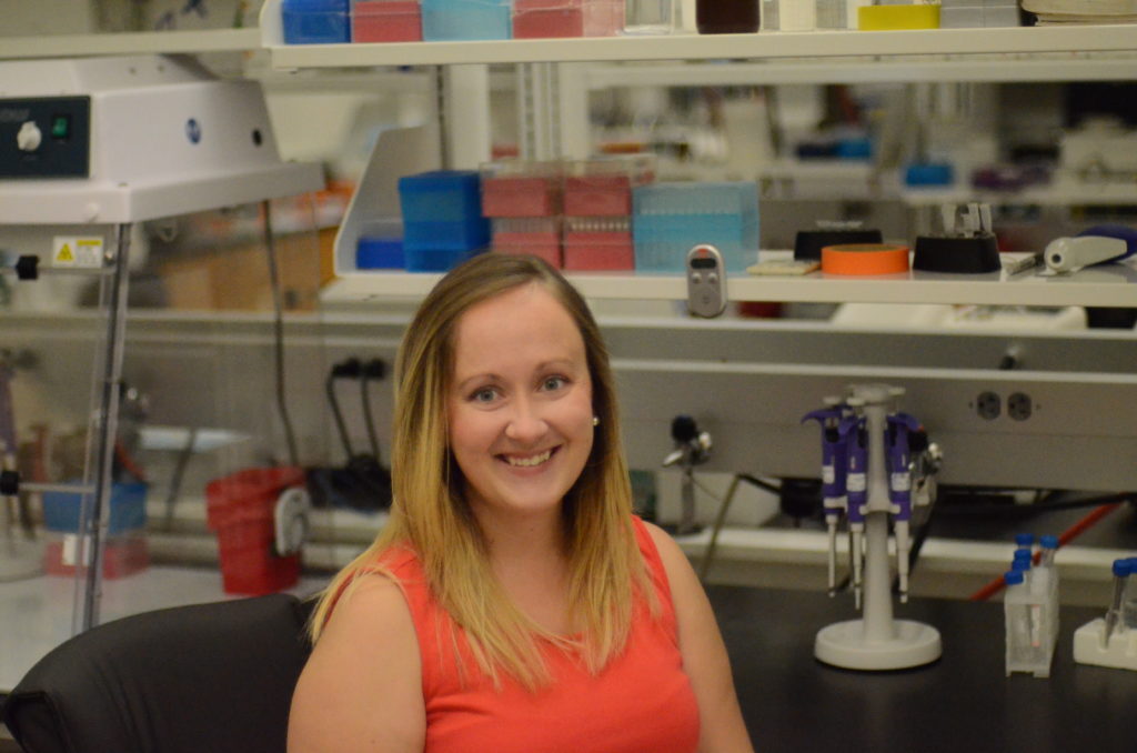 Woman seated at a lab bench in formal clothes smiling.