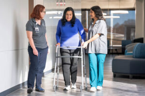 Two nurses assist a patient using a walker.