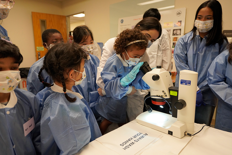A group of children in blue lab coats and masks watches as a young girl looks through a microscope, guided by an adult, in a classroom setting.