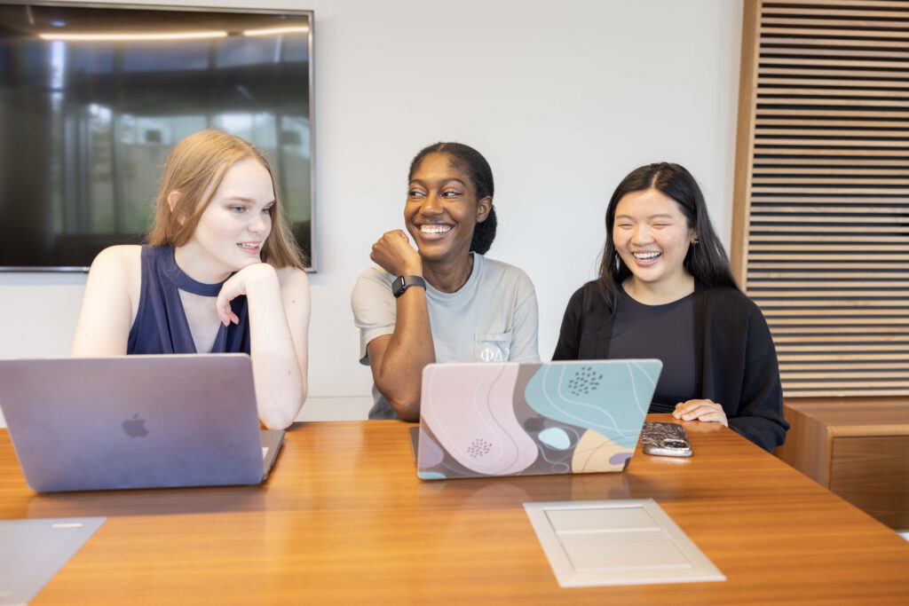Photograph of three students at a table working on laptops and laughing